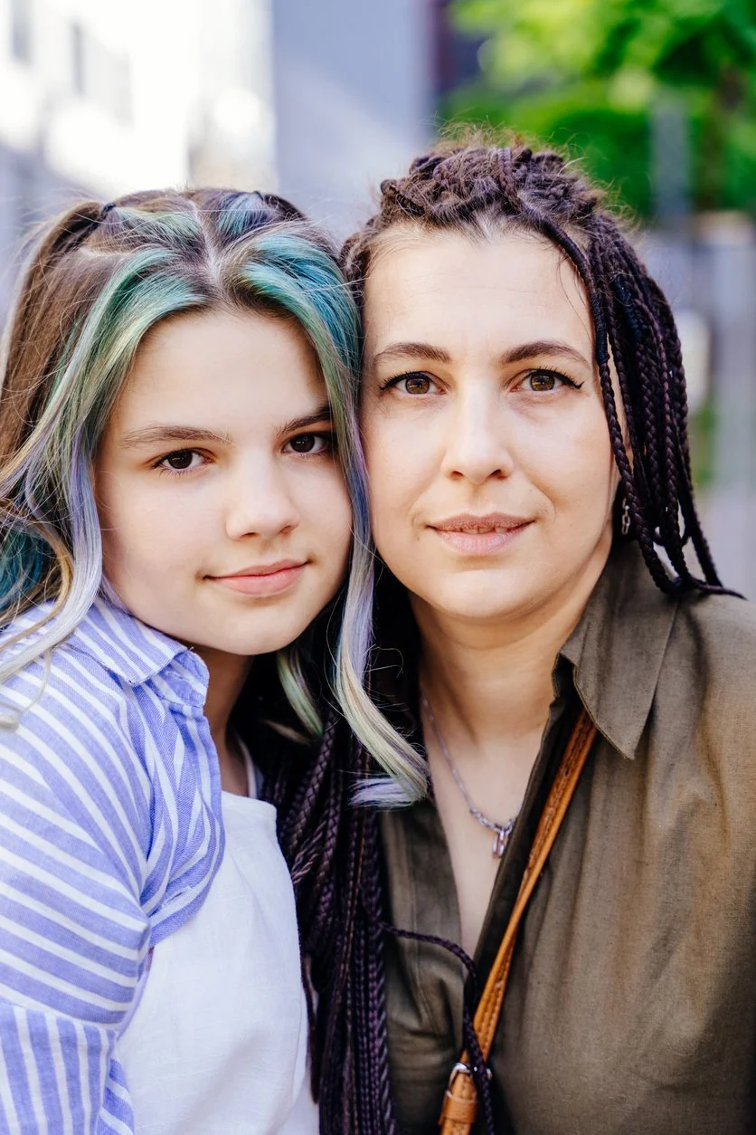 A woman with dark braided hair and a young girl with long multicolored hair close together outdoors.