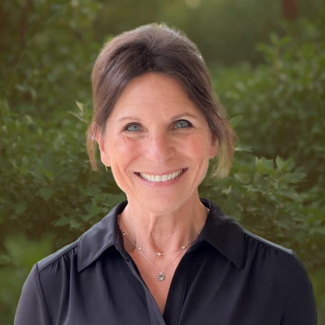 A smiling woman with shoulder-length brunette hair outdoors, wearing a black collared shirt and a delicate necklace, with green foliage in the background.