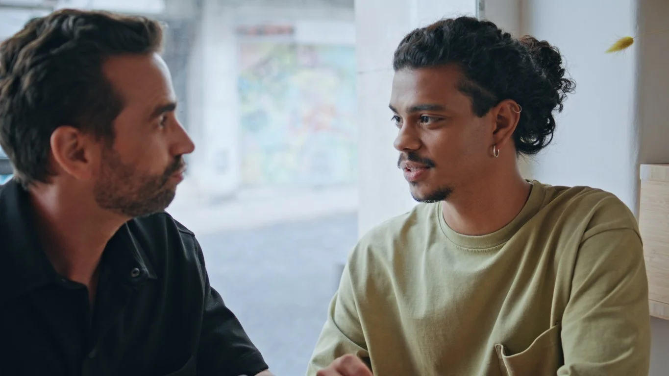 Two men having a conversation in a cozy cafe, sitting close to each other by a window with natural light.