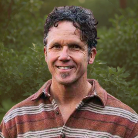 Headshot of a man with curly dark hair smiling outdoors with green foliage in the background, wearing a striped button-up shirt.