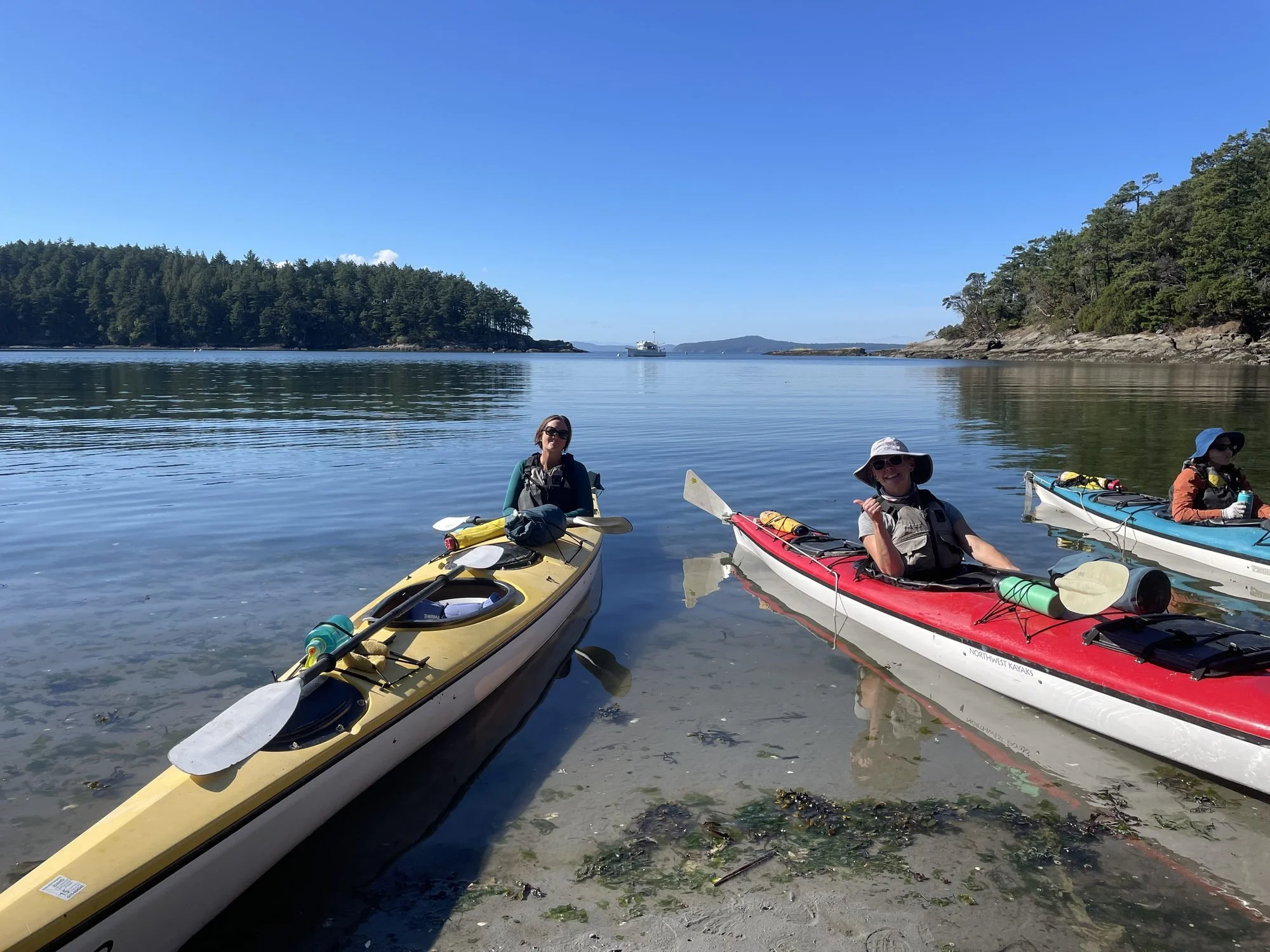 Three people in kayaks on a calm body of water near a forested shoreline with a boat in the background.
