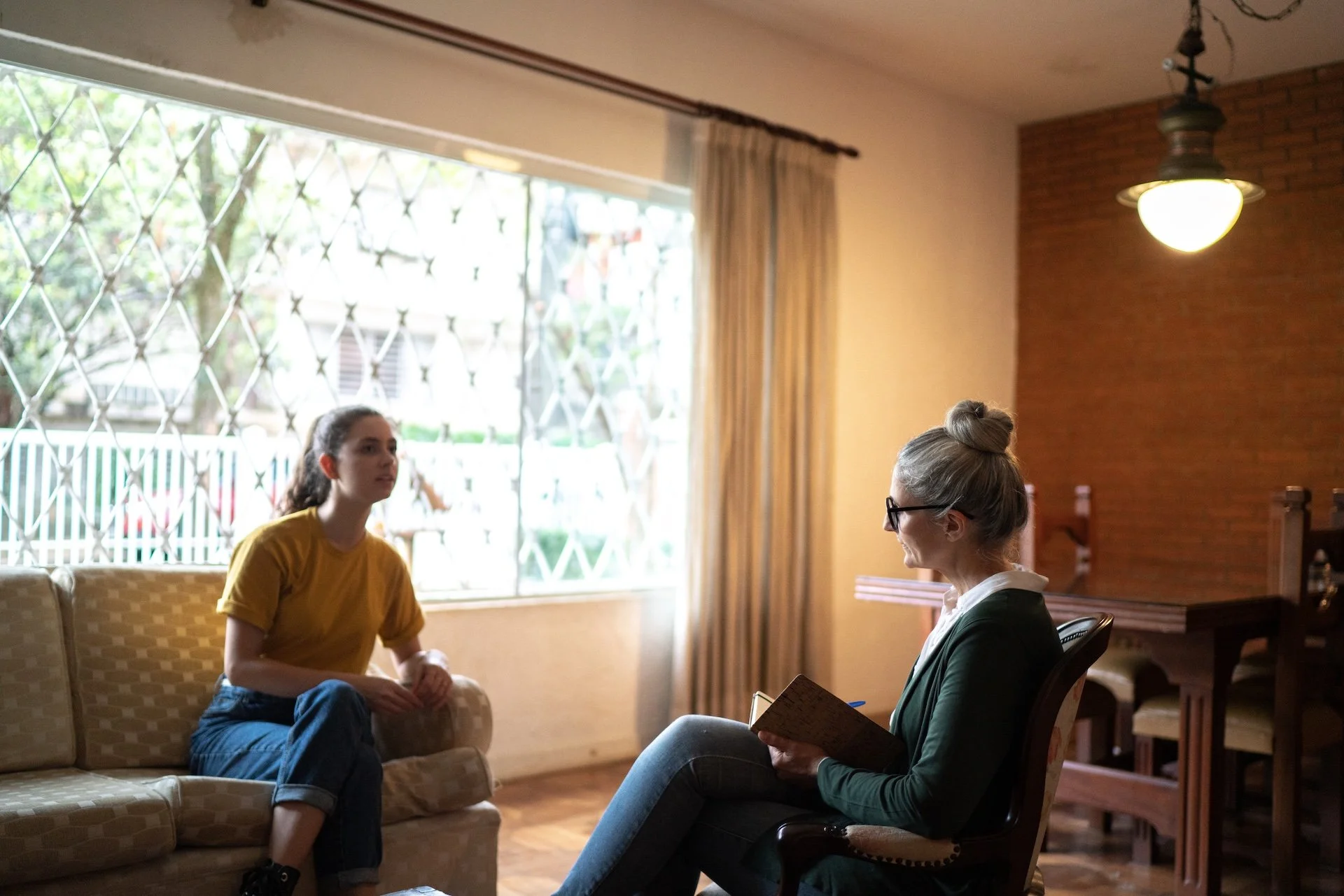 A woman with gray hair and glasses takes notes while talking to a young woman with dark hair in a yellow shirt, sitting on a beige couch in a warmly lit room with a large window and curtains.