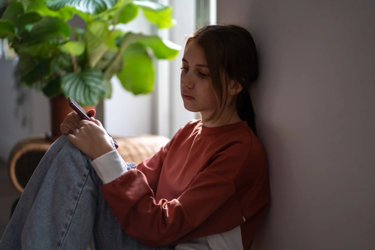 Young woman sitting against a wall, looking at her phone in a room with a large green plant in the background.