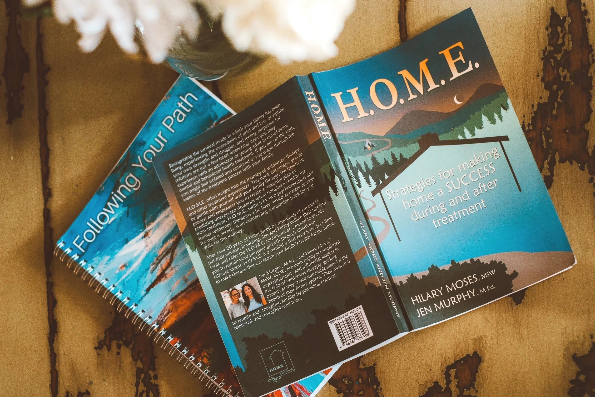 A hardcover book titled 'H.O.M.E.' by Hilary Moses, Jen Murphy, and other authors, with a mountain landscape illustration on the cover, and another book titled 'Follow Your Path' partially visible underneath, placed on a rustic wooden surface.