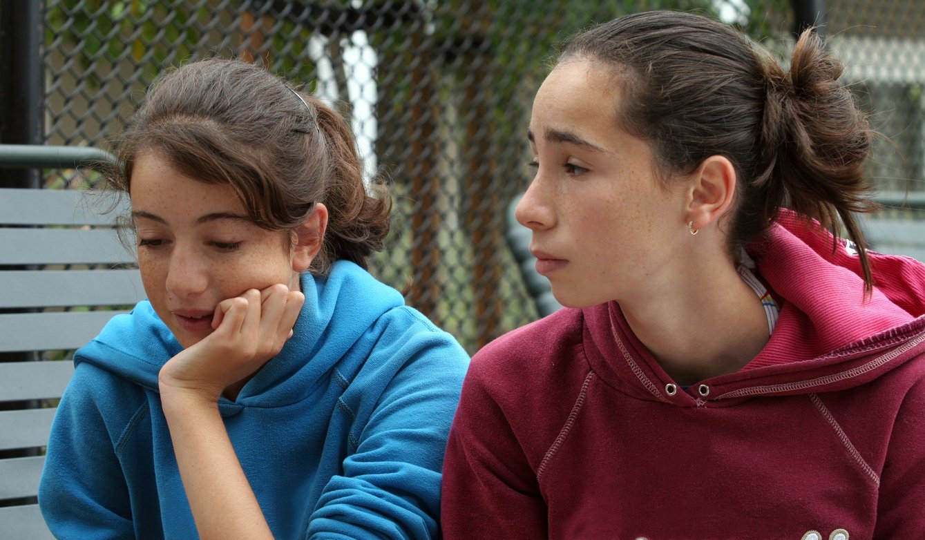Two young women sitting on a park bench, engaged in conversation. The girl on the left is wearing a blue hoodie and resting her face on her hand, while the girl on the right is wearing a magenta hoodie and looking at her friend.