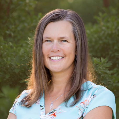 A woman smiling outdoors with green trees in the background, wearing a light blue top with a floral pattern.