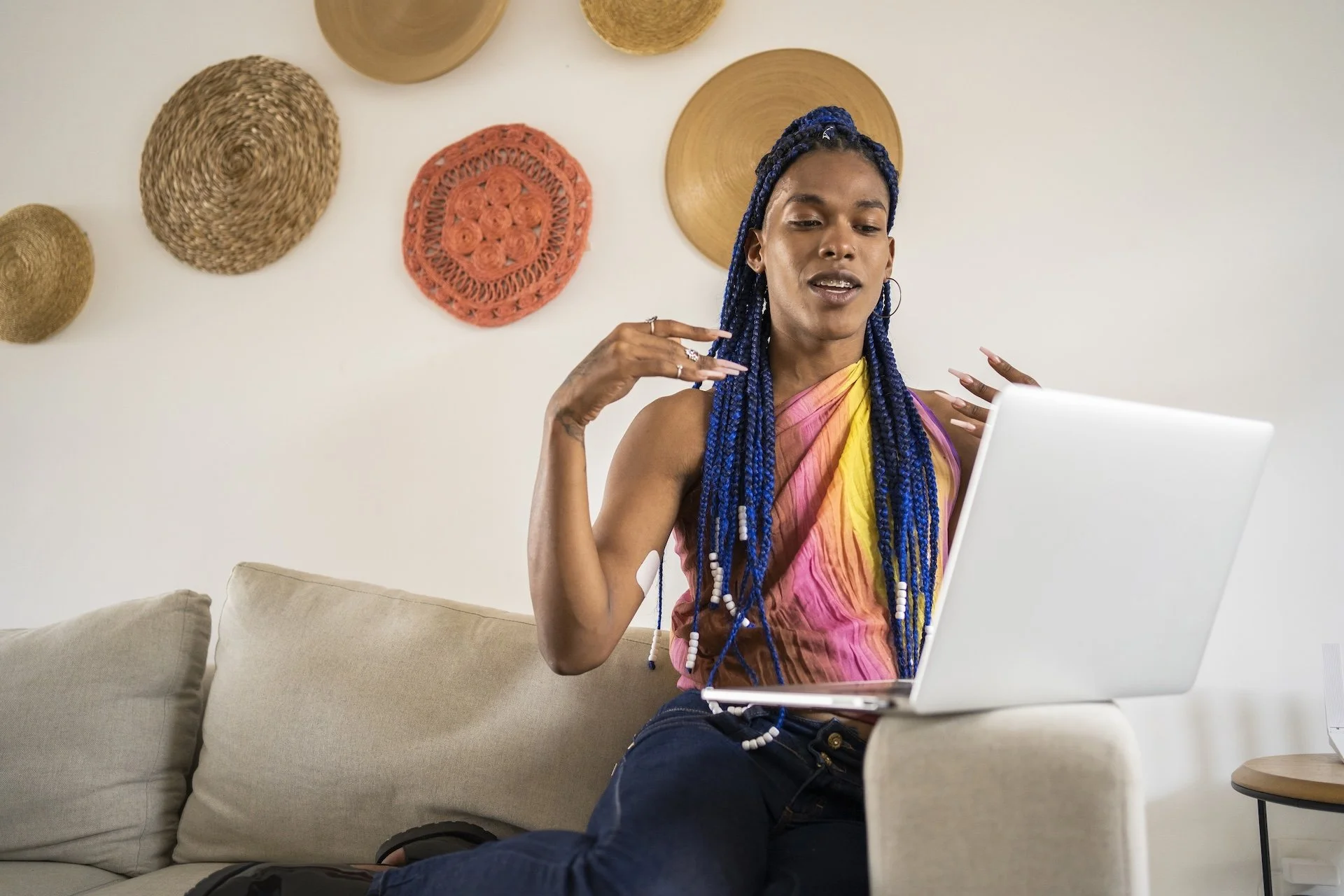 A woman with blue braided hair sitting on a beige sofa, looking at a laptop, gesturing with her right hand, in a room with woven wall decorations.