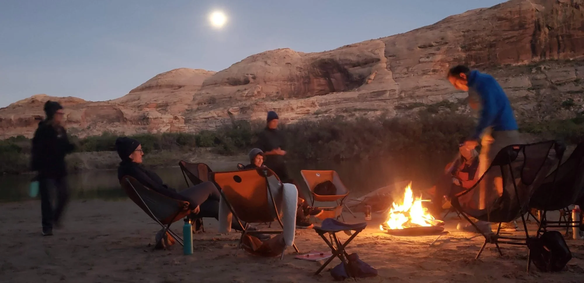 Group of people sitting and standing around a campfire near a river with desert cliffs in the background during dusk.