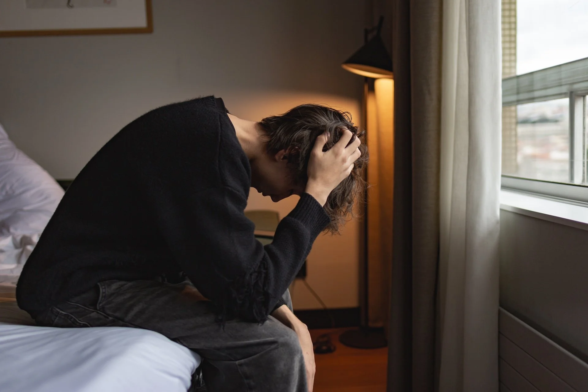 Person sitting on bed holding their head with both hands in a dimly lit room near a window.