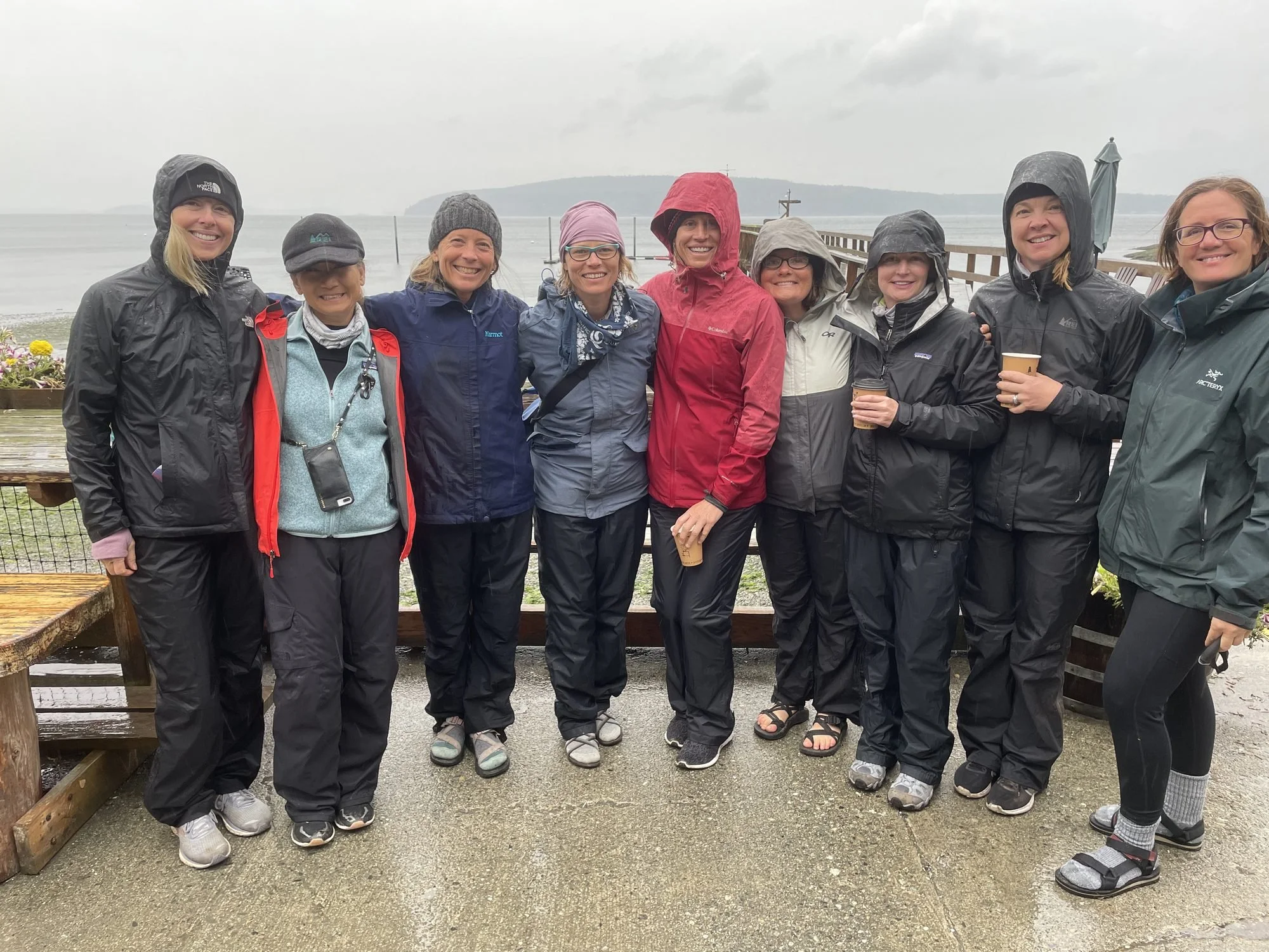 A group of ten women standing outdoors on a rainy day with the ocean and a distant landmass in the background. They are dressed in rain jackets, hats, and waterproof shoes, smiling and holding coffee cups.