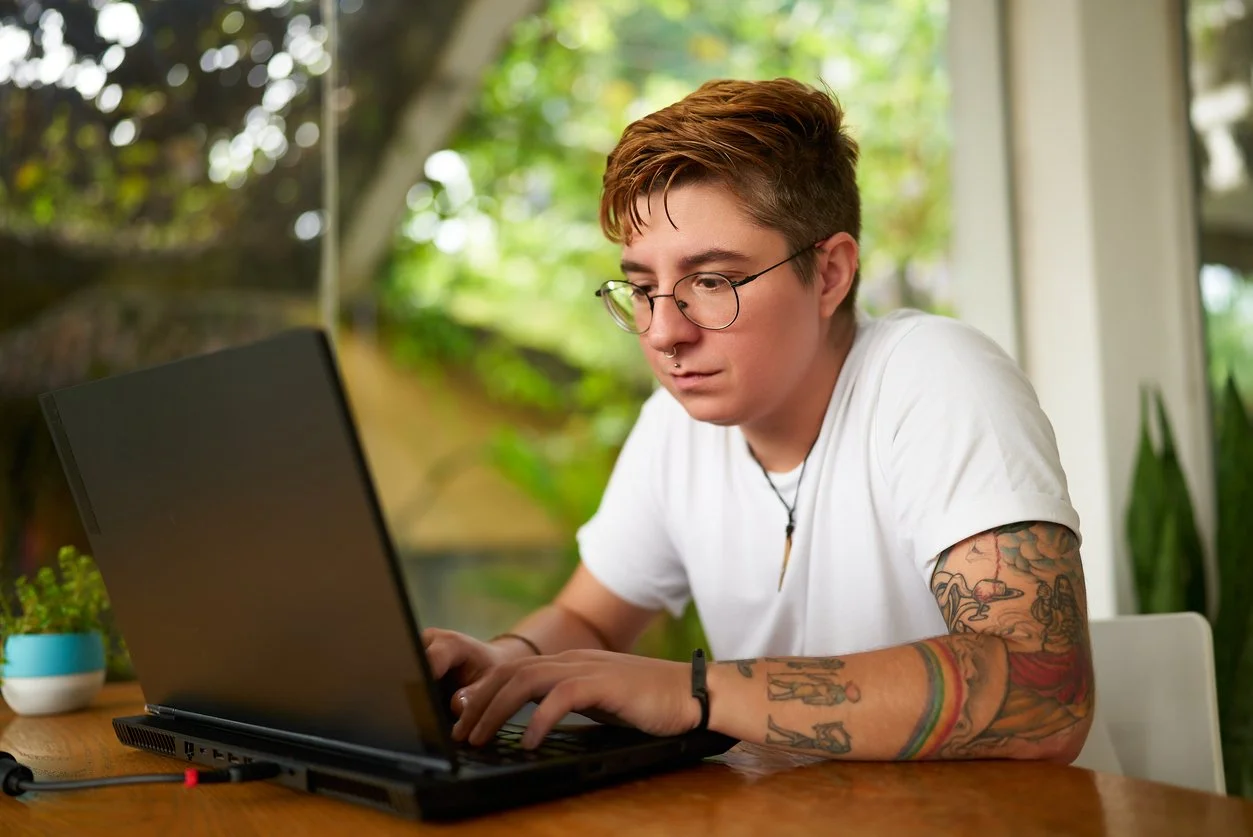 Person with short hair and glasses working on a laptop, sitting at a wooden table with a potted plant nearby, in a bright room with large windows and greenery outside.