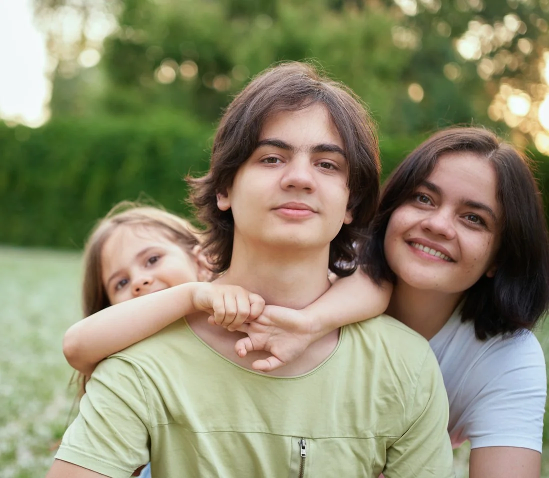 A family of three outdoors, with the teenage son in front, a woman behind him, and a young girl on his shoulder, all smiling.