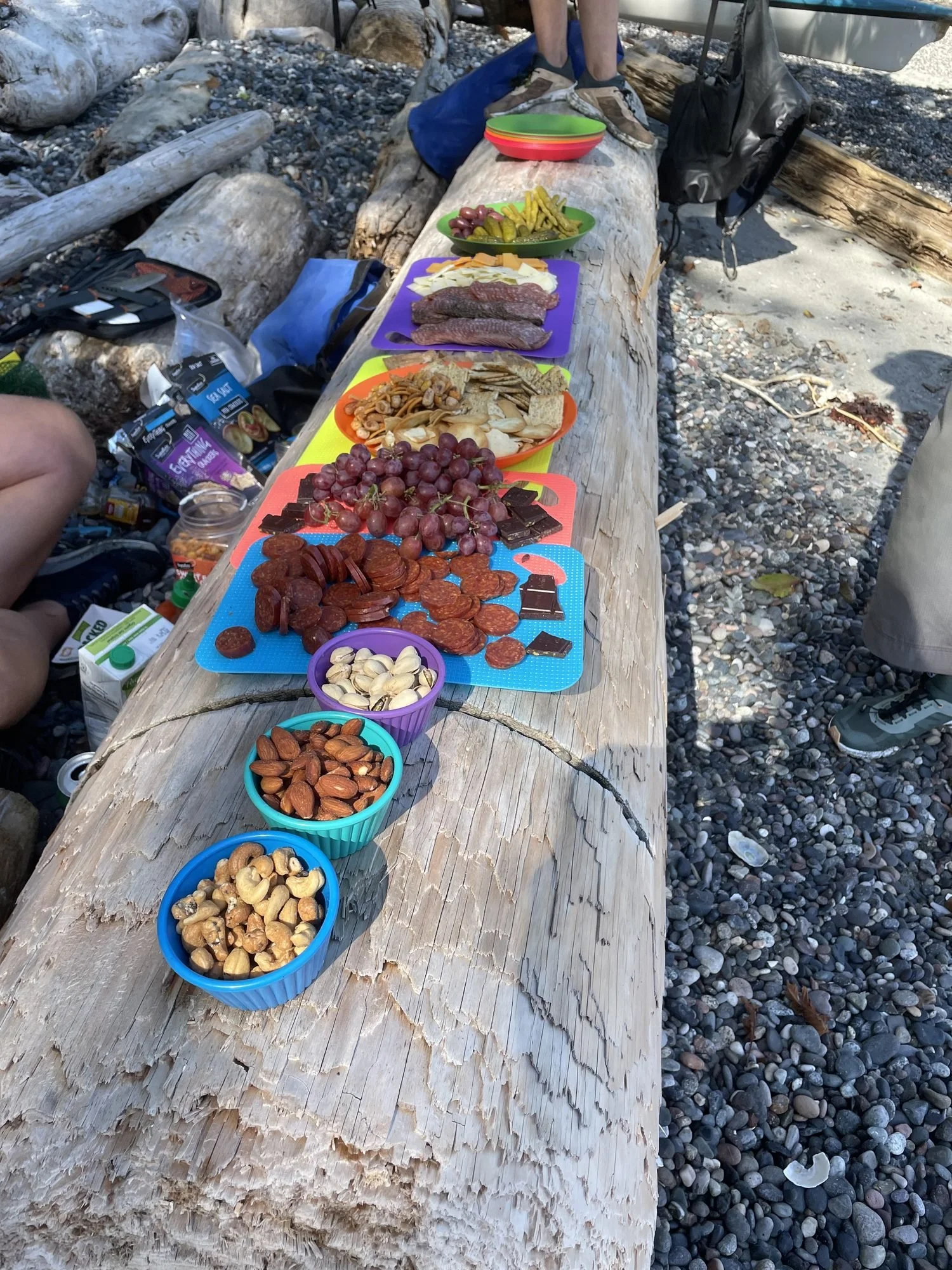 A makeshift outdoor food setup on a log with various plates of snacks and food items including grapes, sliced meats, cheese, crackers, and mixed nuts on colorful trays. Some camping gear and people are visible in the background on a beach with pebbles.