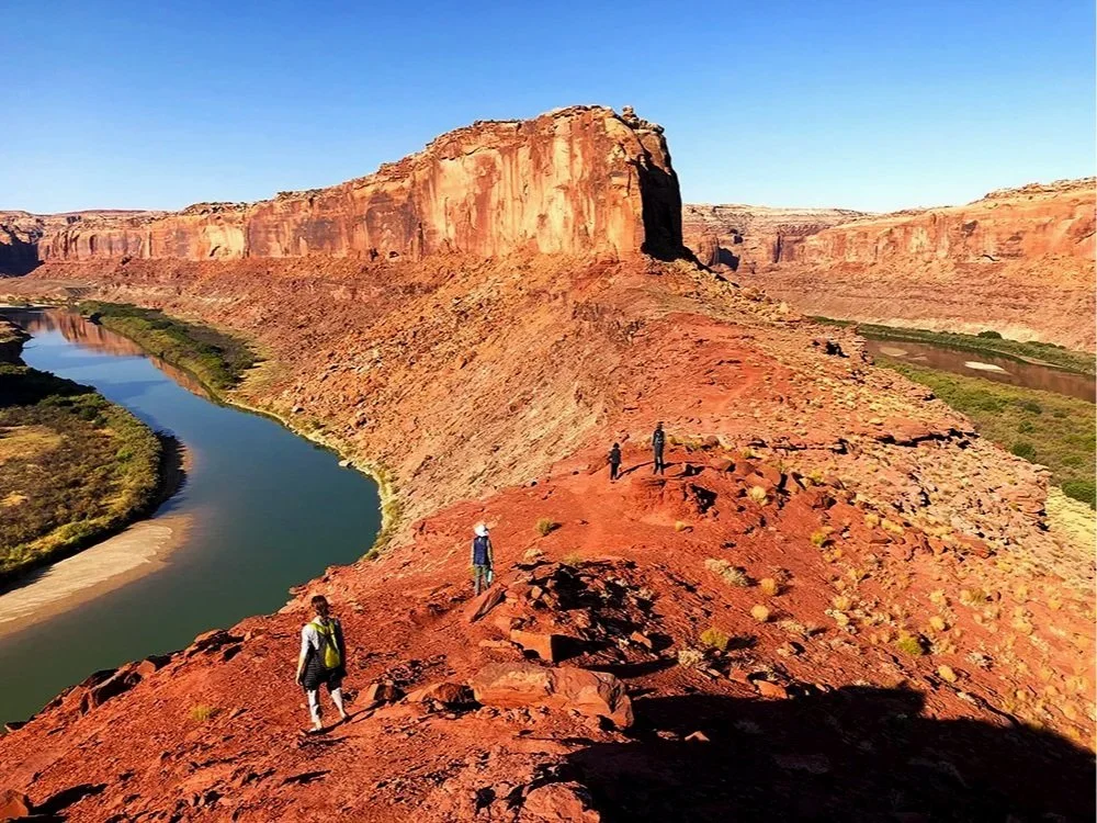 Hikers walking on a rocky red desert trail near a winding river with steep canyon walls under a clear blue sky.