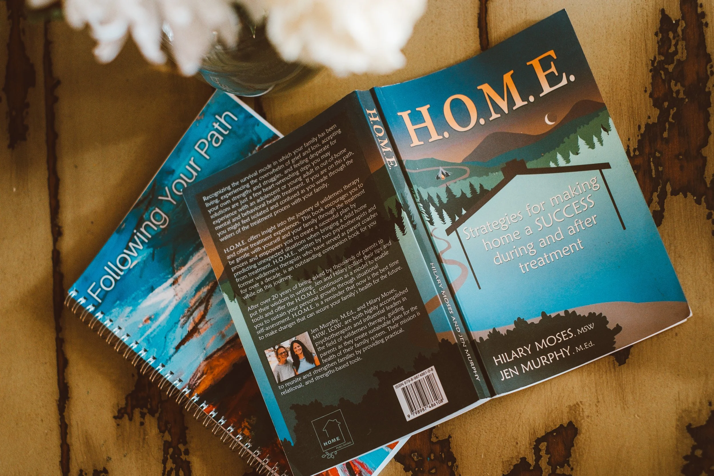 A collection of books on a wooden table, including one titled 'H.O.M.E.' about strategies for making home a success. The books have colorful covers and are accompanied by a small part of a flower in a glass container.