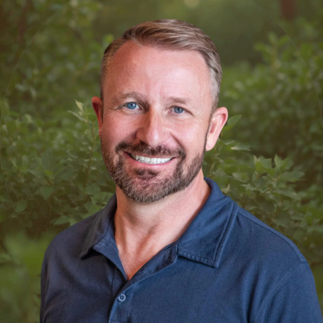A man with short, light brown hair and a beard, smiling in front of green foliage, wearing a navy blue collared shirt.