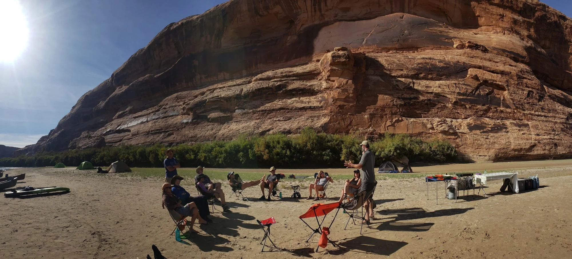 A group of people sitting in camping chairs on a sandy beach, with a large rocky cliff in the background and camping tents nearby, sunny weather.