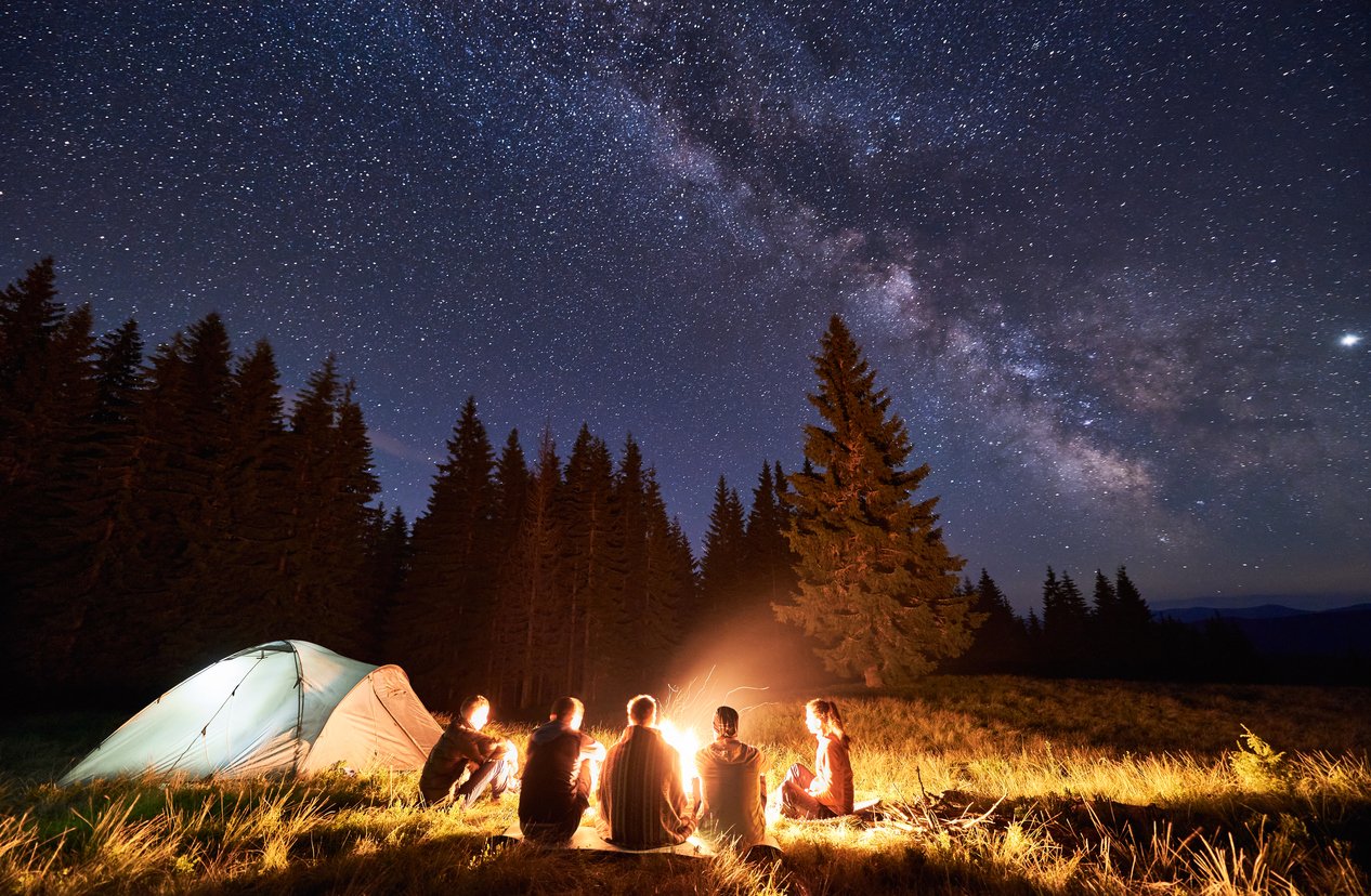 Five people sitting around a campfire at night in a grassy field surrounded by trees, with a bright starry sky and the Milky Way visible overhead, and a tent nearby.
