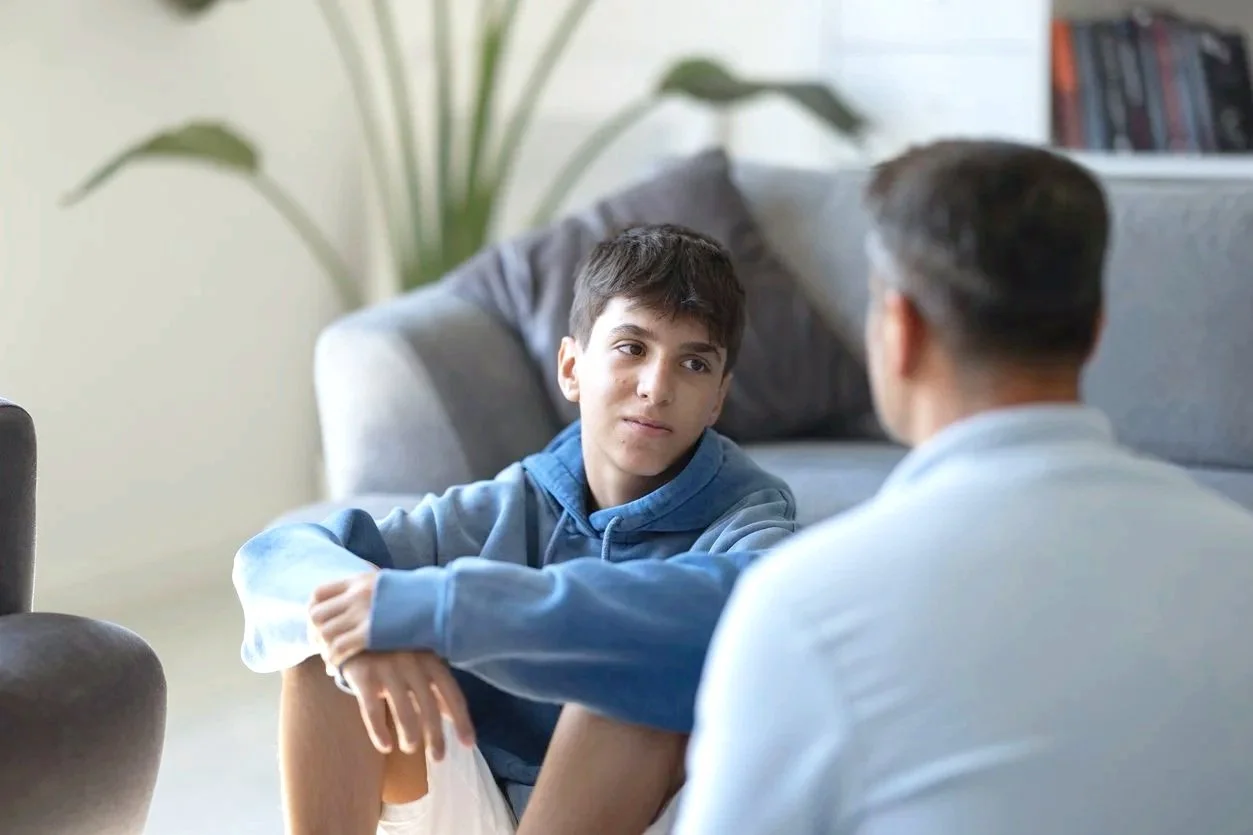 A young boy sitting on the floor looking at a man, possibly a therapist, during a session in a living room. The boy is wearing a hoodie and shorts, and the man has short hair and glasses.