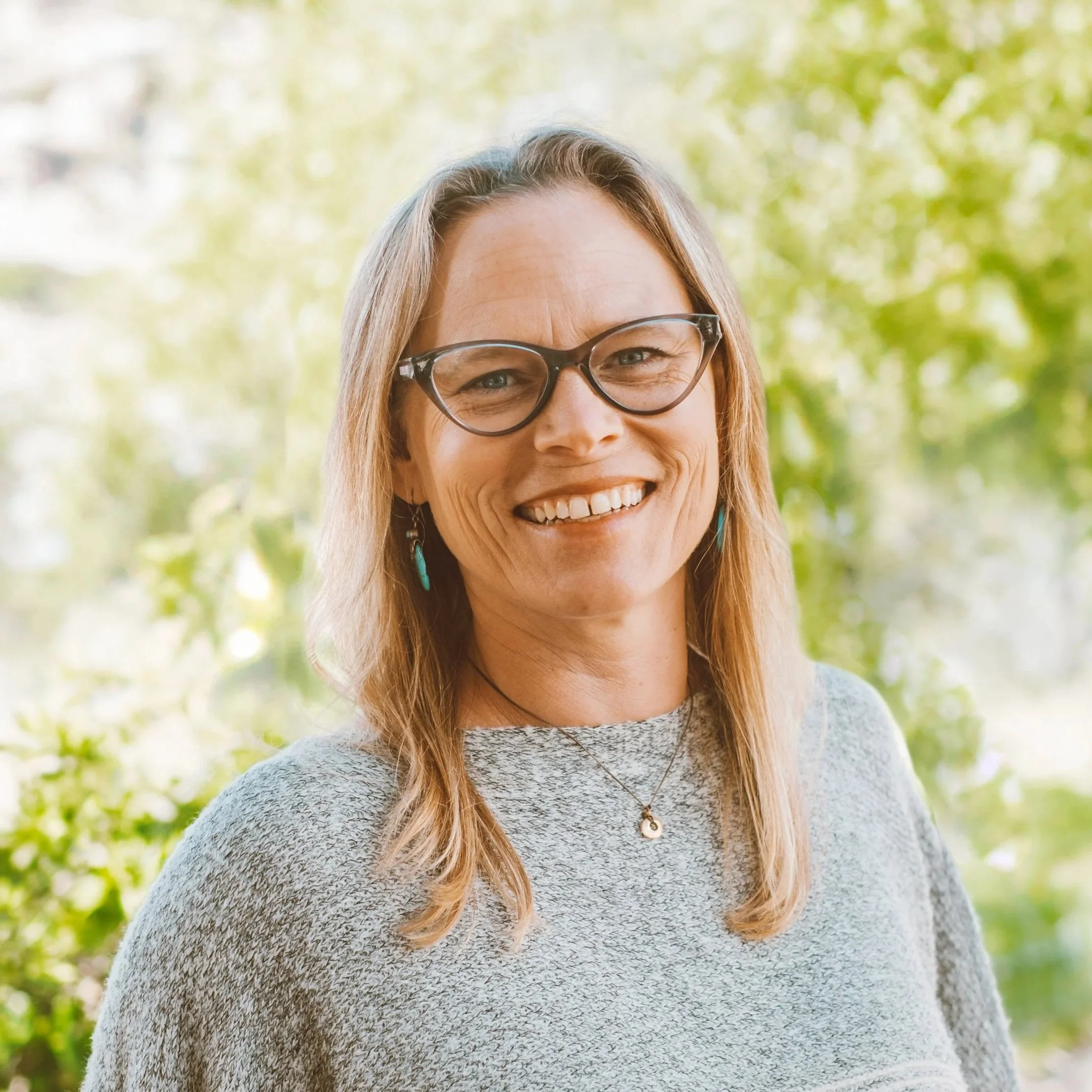 A woman with long blond hair, wearing glasses and a gray patterned sweater, smiling outdoors with blurred green trees in the background.