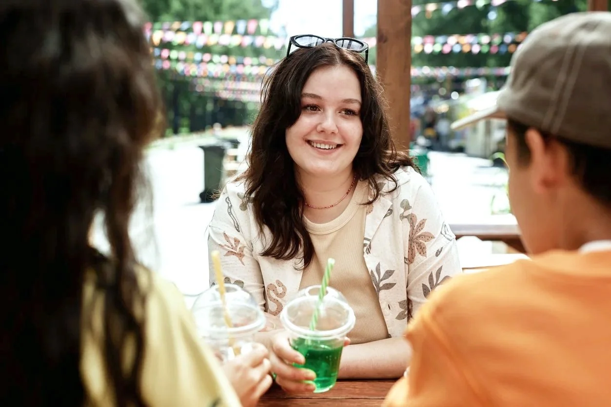 A woman with dark wavy hair and glasses on her head smiles while talking with two children at an outdoor table, each holding a cup of bright green drink with a straw. In the background, colorful string lights are hung outdoors.