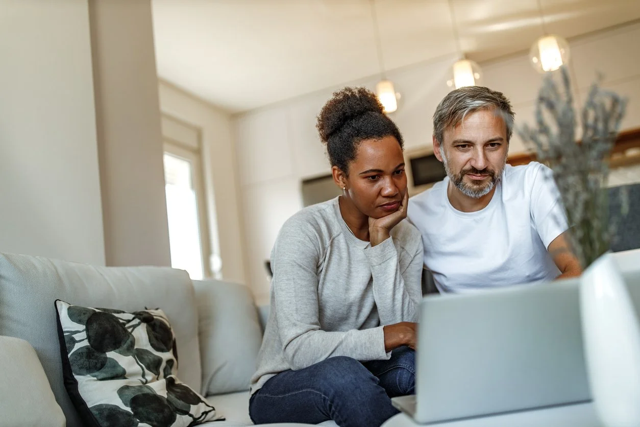A man and woman looking at a laptop screen together on a beige couch in a living room.