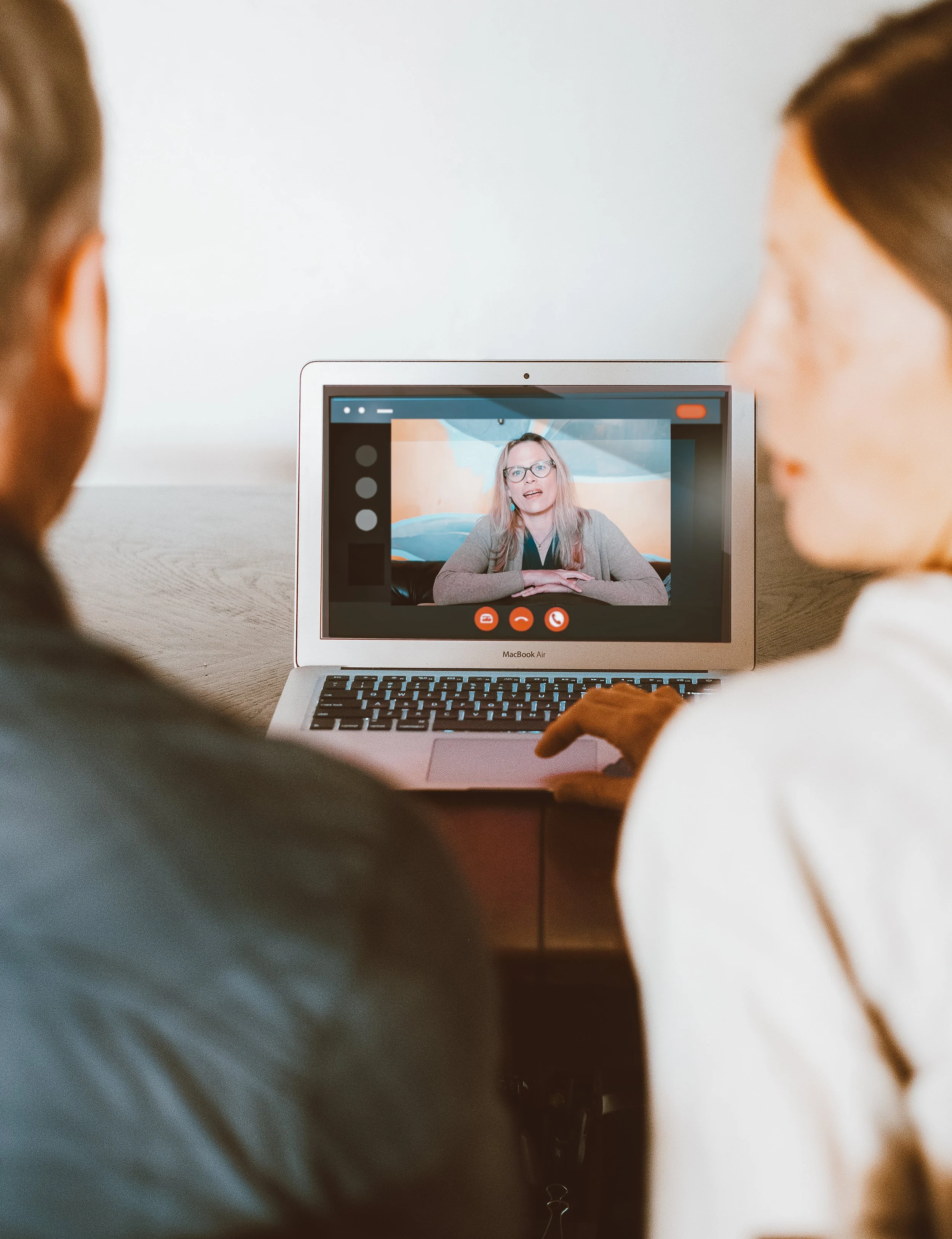 People are watching a woman speak on a video call displayed on a laptop.