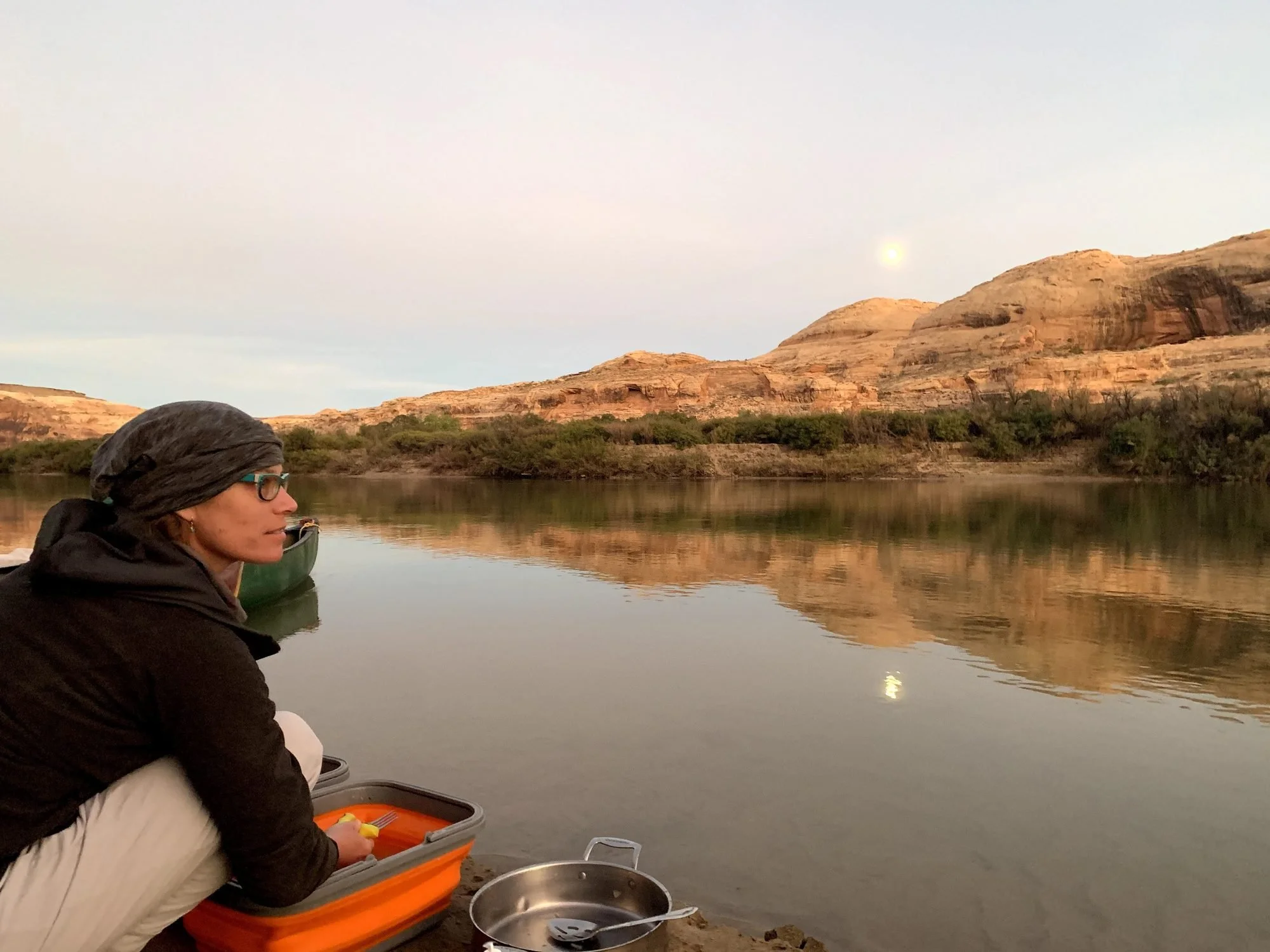 A woman is sitting by a river at sunset, preparing food from a lunchbox, with rocks and hills in the background.