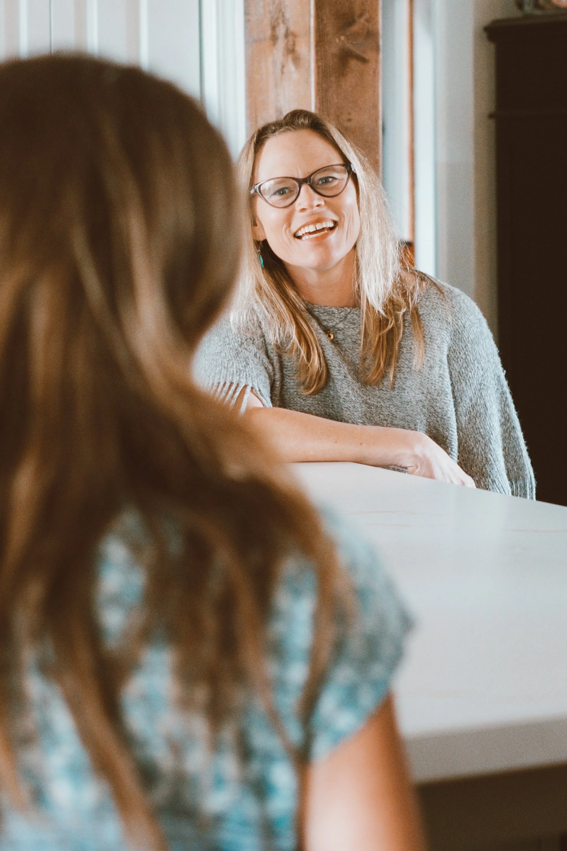 Woman with glasses smiling, sitting at a table, in focus. Blurred person in the foreground.