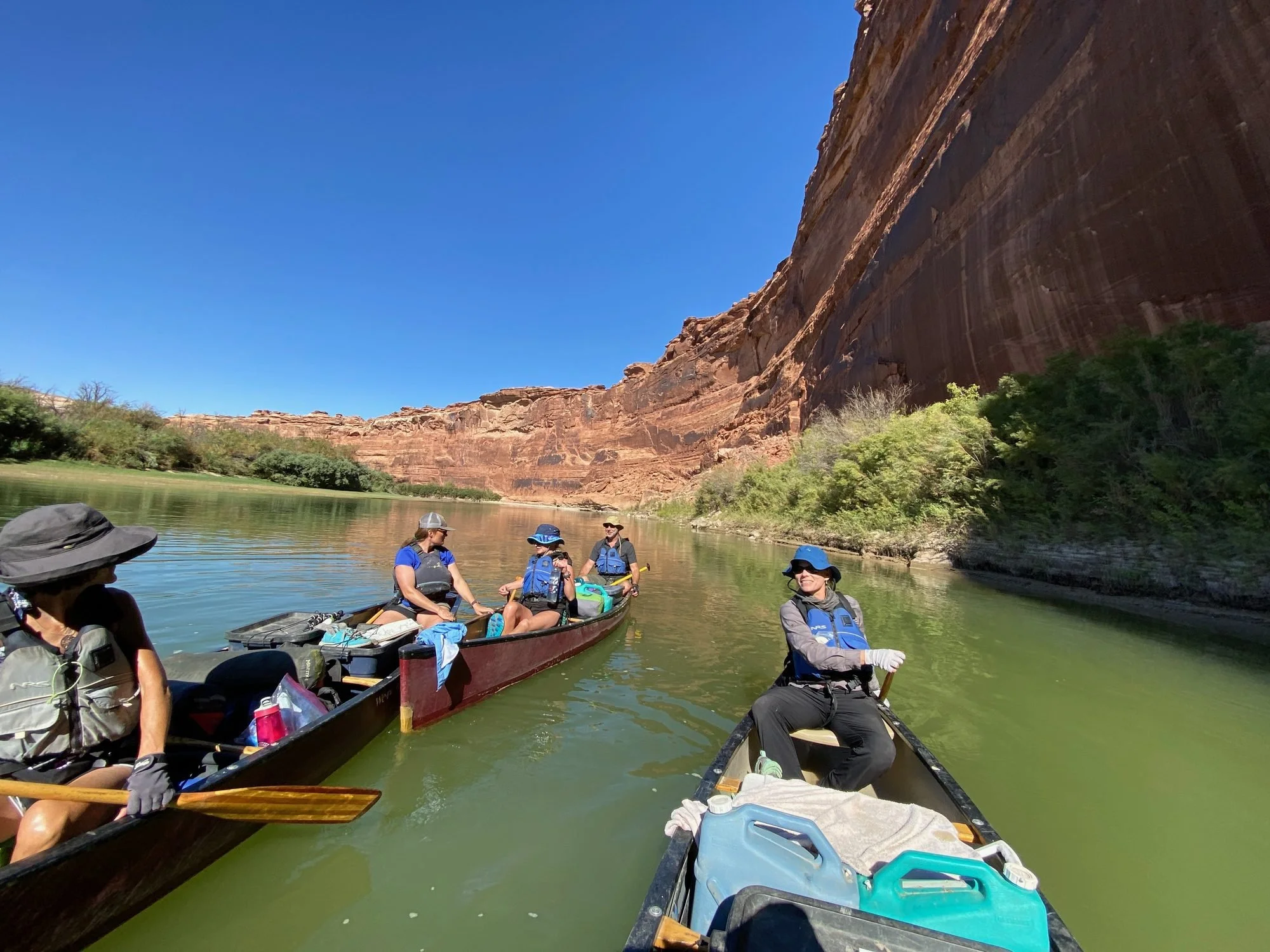 People kayaking on a river with red canyon walls and clear blue sky in the background.