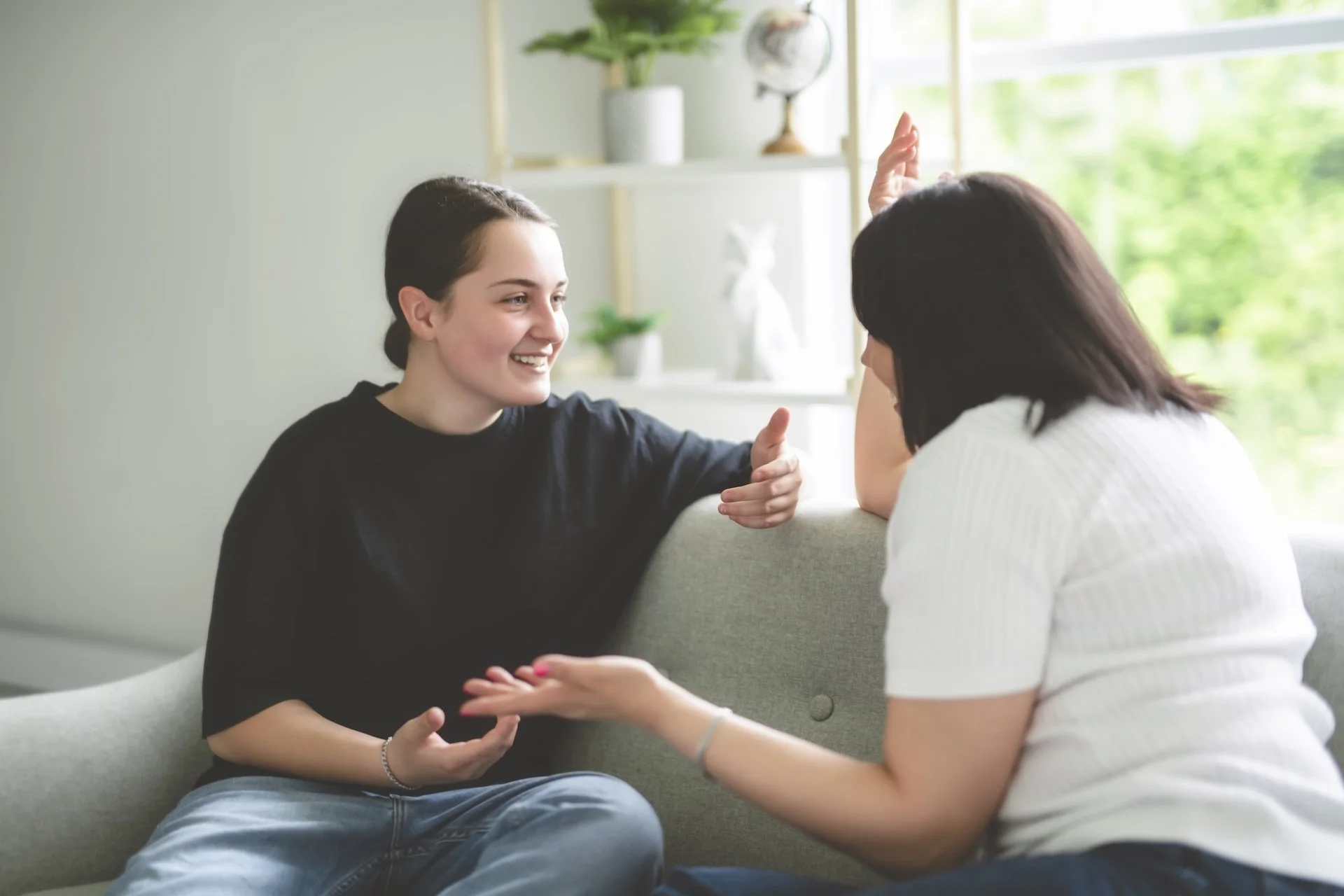 Two women are engaged in a conversation in a bright, modern living room with large window. One woman with dark hair in a ponytail, wearing a black shirt, is smiling and gesturing with her hands. The other woman, with dark hair and a white shirt, is talking and sitting on the sofa.