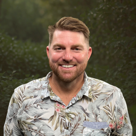 A man with short brown hair and a beard smiling outdoors, wearing a floral button-up shirt.