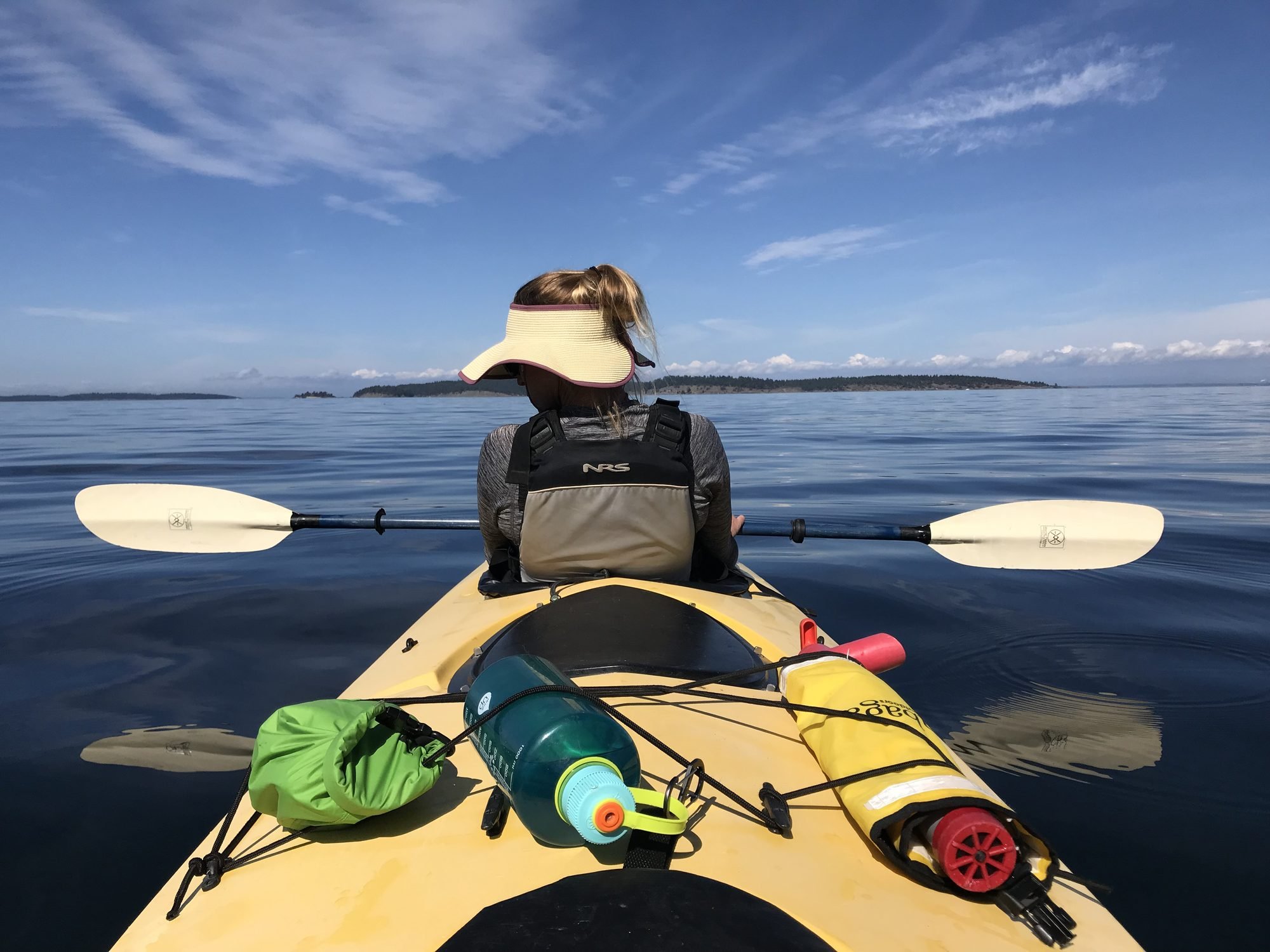 Person kayaking on a calm body of water with a wide blue sky and distant landforms in the background, carrying gear including a water bottle, paddle, and safety equipment.