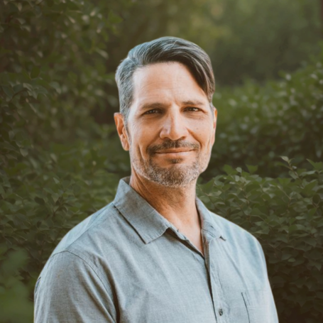A middle-aged man with gray hair and a beard, wearing a light gray collared shirt, standing outdoors in front of greenery, smiling slightly.