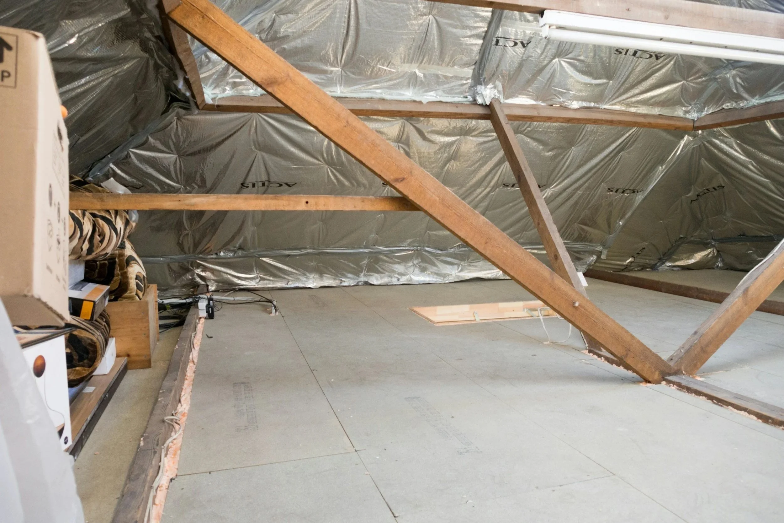 Une pièce sous les combles en construction avec des poutres en bois et une isolation en aluminium au plafond, la moitié inférieure du mur est en train d'être rénovée avec des matériaux de construction.