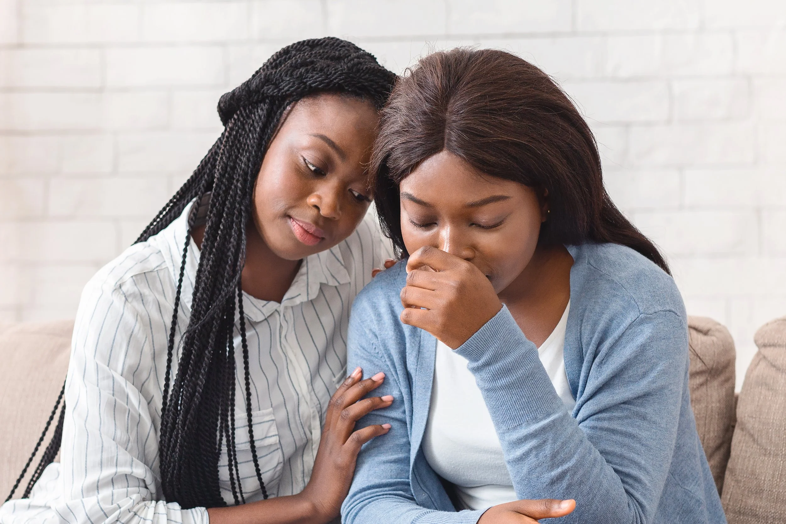 Two women comforting each other; one woman appears distressed while the other offers support, sitting on a beige couch with a white brick wall background.