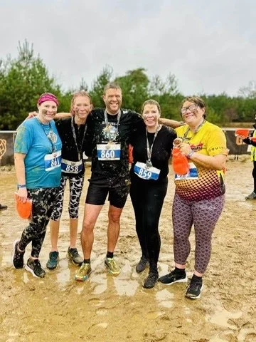 Outdoor fitness in Cranleigh. Group of five people dressed in athletic clothing, posing together outdoors after a race, some wearing bib numbers, with trees and a muddy ground in the background.