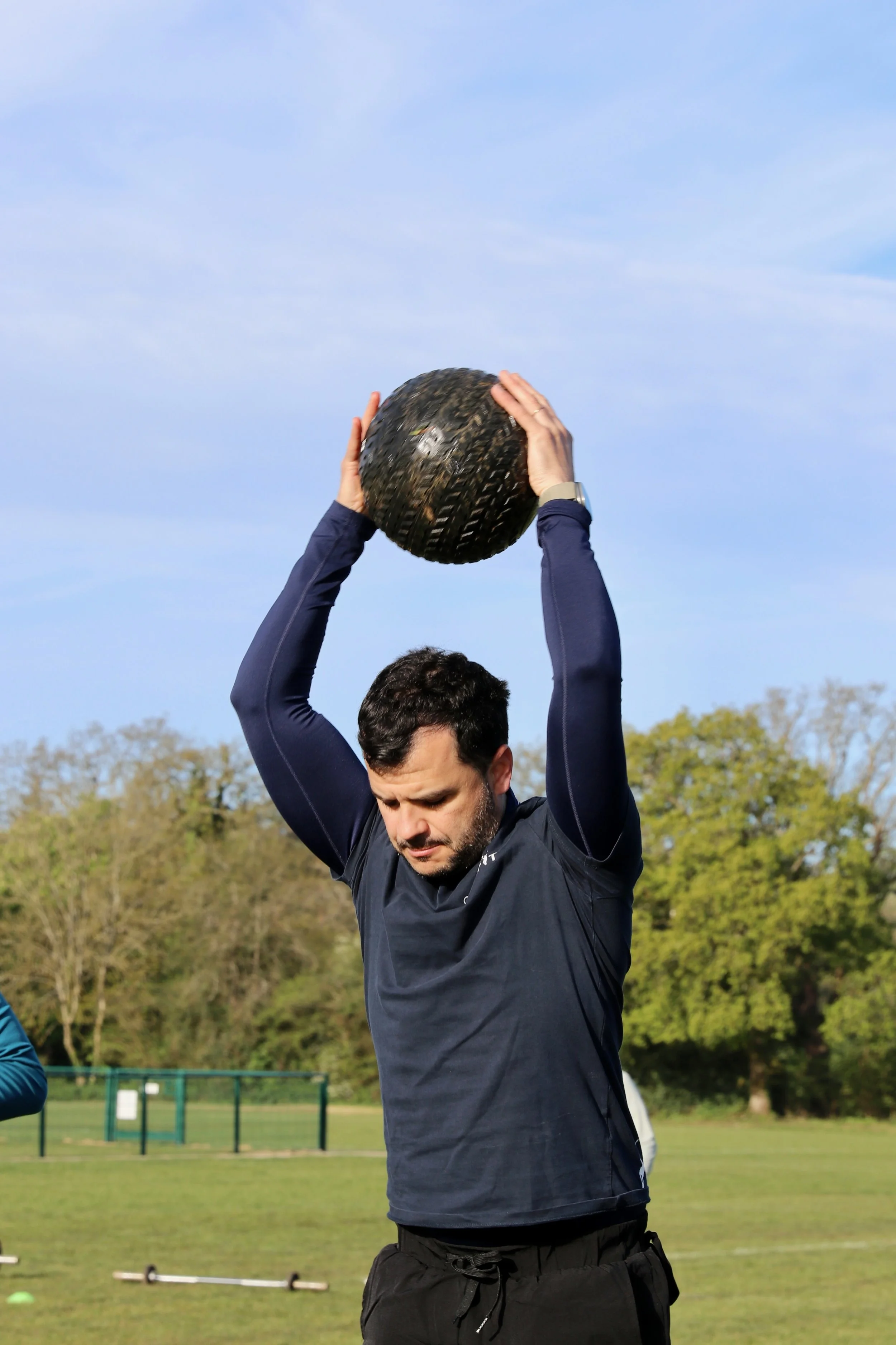 A man holding a black medicine ball above his head outdoors on a grassy field with trees in the background.