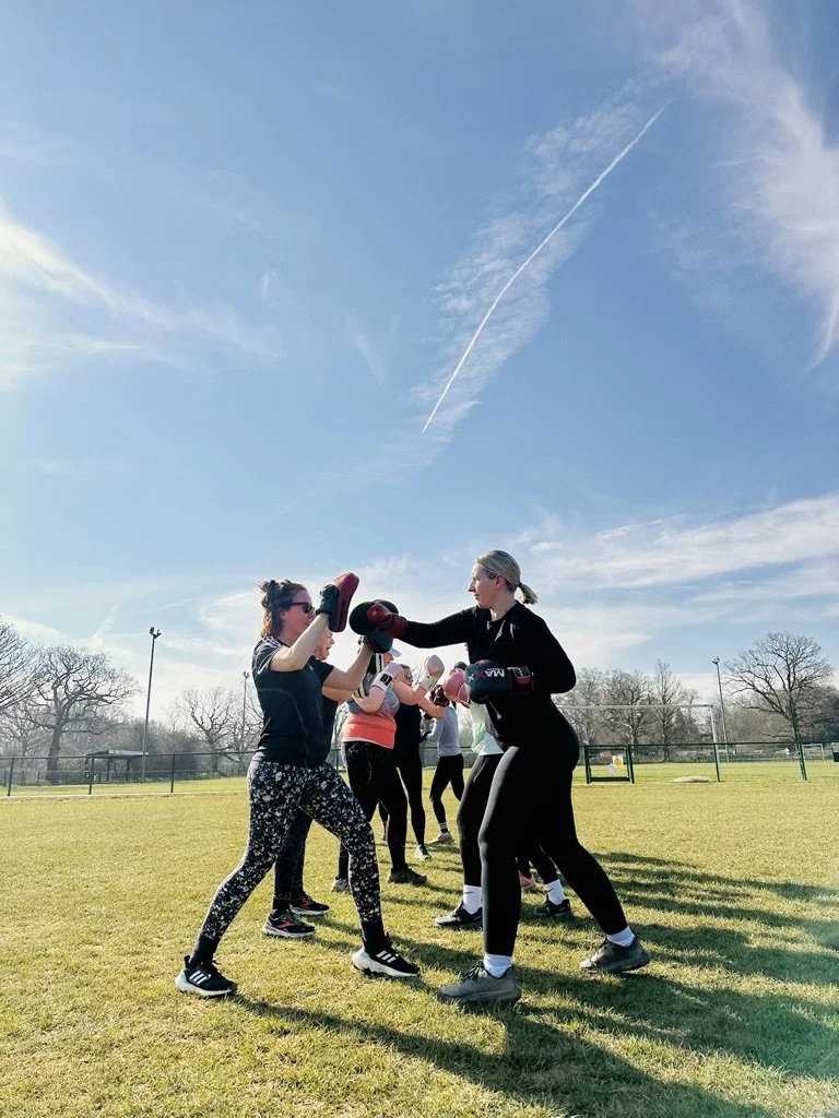 Outdoor fitness in Cranleigh. Women participating in a boxing class outdoors on a grassy field with women holding focus mitts and wearing boxing gloves, under a blue sky with a contrail.