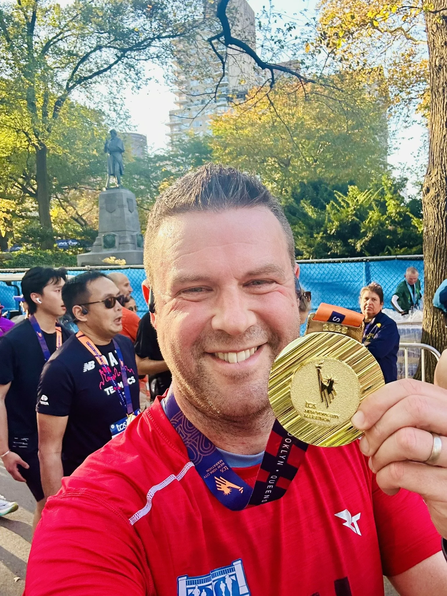 A man smiling and holding a gold marathon medal at a race event, with other runners and a park with trees and a statue in the background.