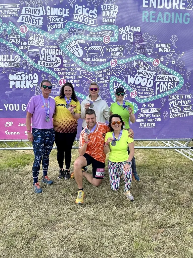 Outdoor fitness in Cranleigh. Group of five people, three women and one man, wearing medals and athletic gear, posing in front of a large colorful illustrated map at a race or marathon event. The map features various trails and motivational phrases.