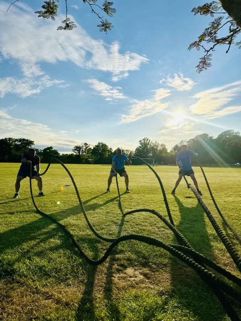 Outdoor fitness in Cranleigh. Three people exercising outdoors in a grassy field during sunset, using battle ropes for strength training.