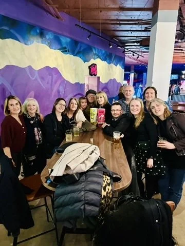Outdoor fitness in Cranleigh. Group of people gathered around a bar table inside a colorful arcade or entertainment venue. They are smiling and holding drinks.
