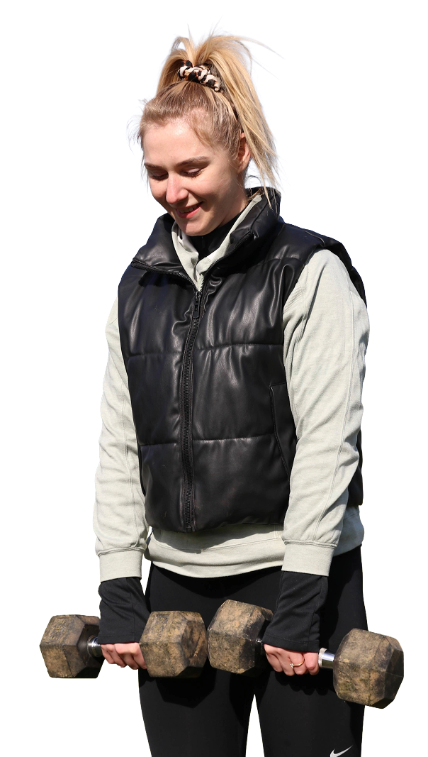 A young woman with blonde hair in a high ponytail, wearing a black vest over a light gray hoodie, and lifting a dumbbell outdoors.