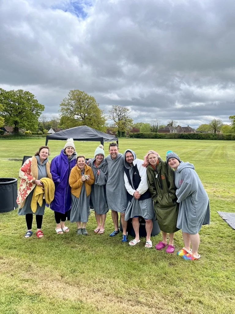 Outdoor fitness in Cranleigh. A group of eight people standing together outdoors on a grassy field, with some wearing rain ponchos and hoodies, under cloudy skies, smiling and posing for a photo.
