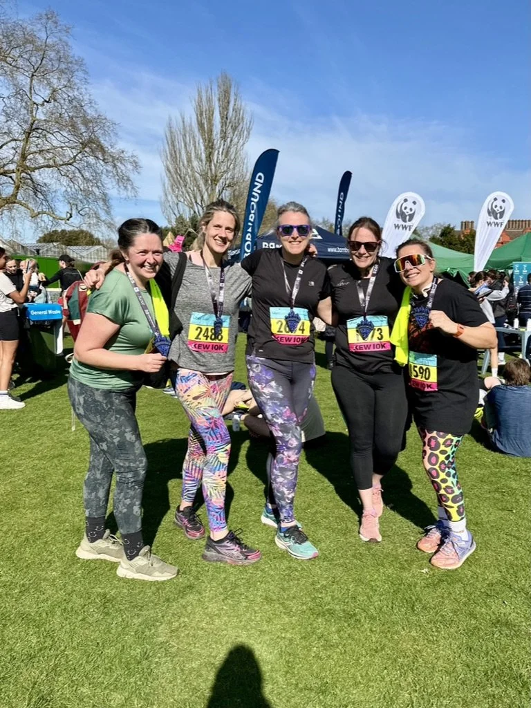 Outdoor fitness in Cranleigh. Five women standing together outdoors at a running event, wearing athletic clothing with race bibs, holding medals, and smiling.
