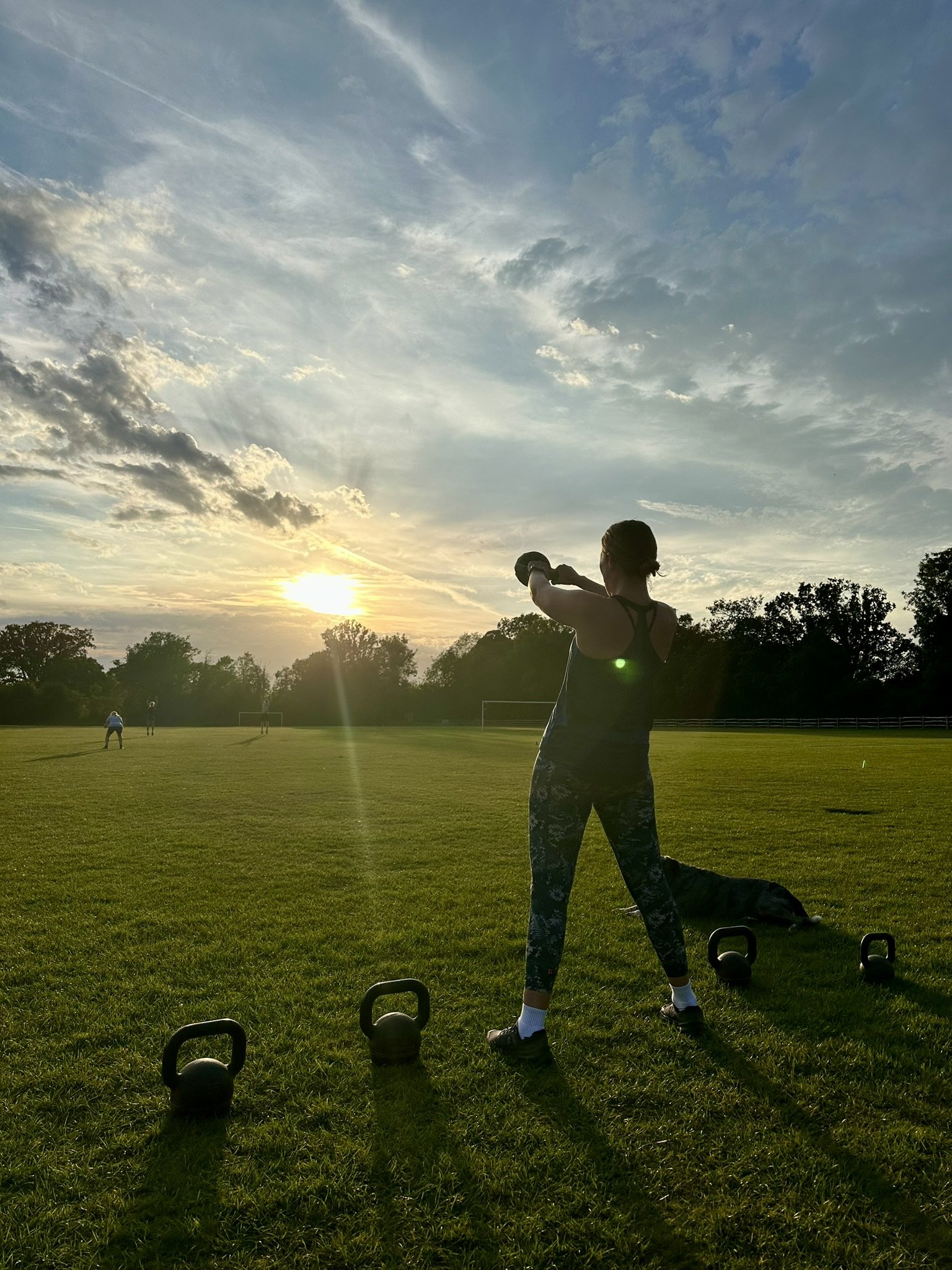 Outdoor fitness in Cranleigh. Person lifting dumbbell during outdoor workout at sunset, with kettlebells on grass, trees, and cloudy sky in background.