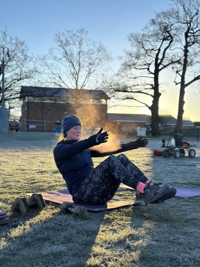Outdoor fitness in Cranleigh. A woman exercising outdoors on a mat at sunrise or sunset in a park, with trees and a building in the background. She is wearing workout clothes, gloves, and a beanie, appears to be stretching or doing a core exercise.