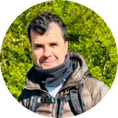 Man outdoors wearing a jacket and scarf, with green trees in the background.