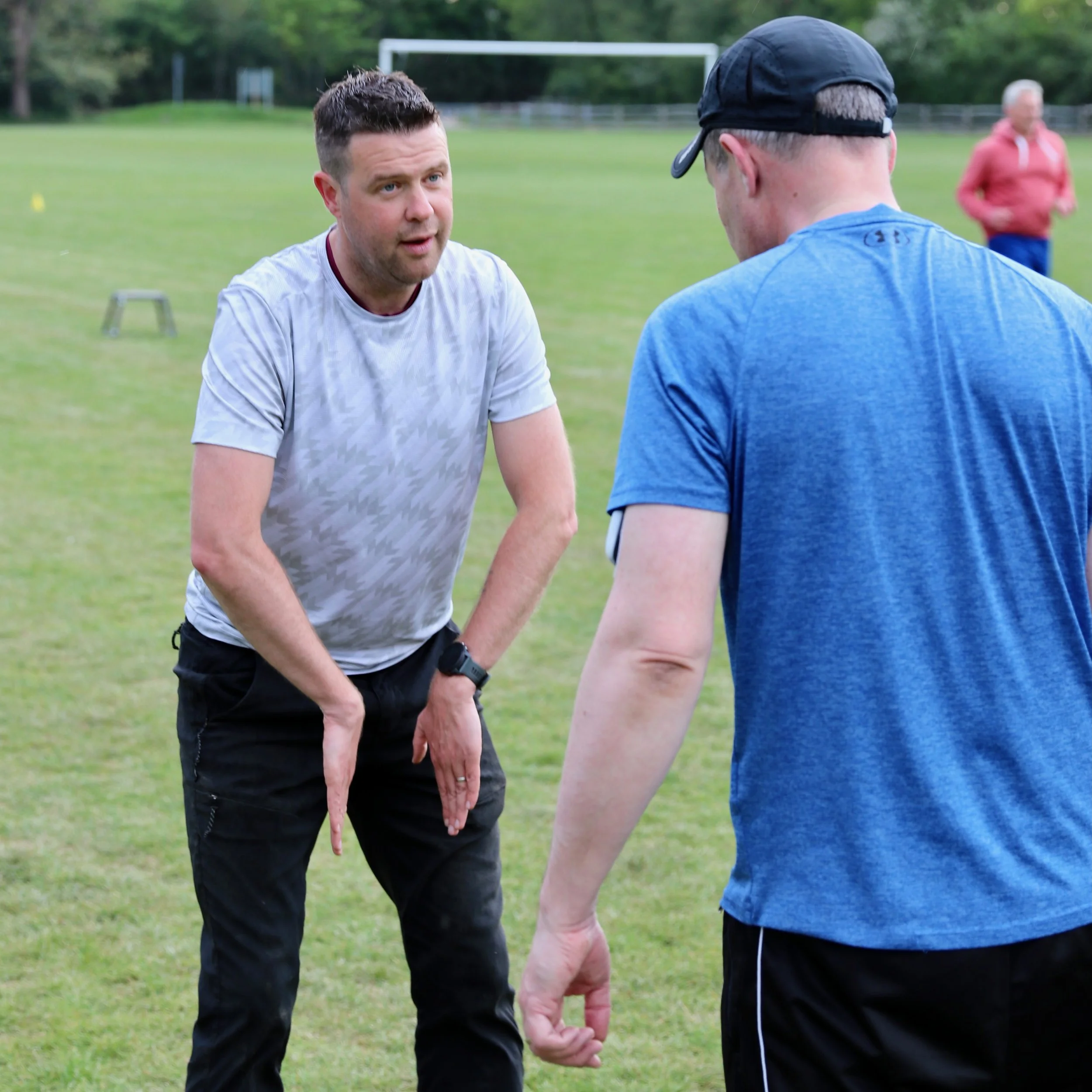 Two men are holding hands in a park with a soccer goal in the background. One man is wearing a white shirt and black pants, and the other man is wearing a blue shirt and a black cap. A woman in a pink jacket is walking in the background.